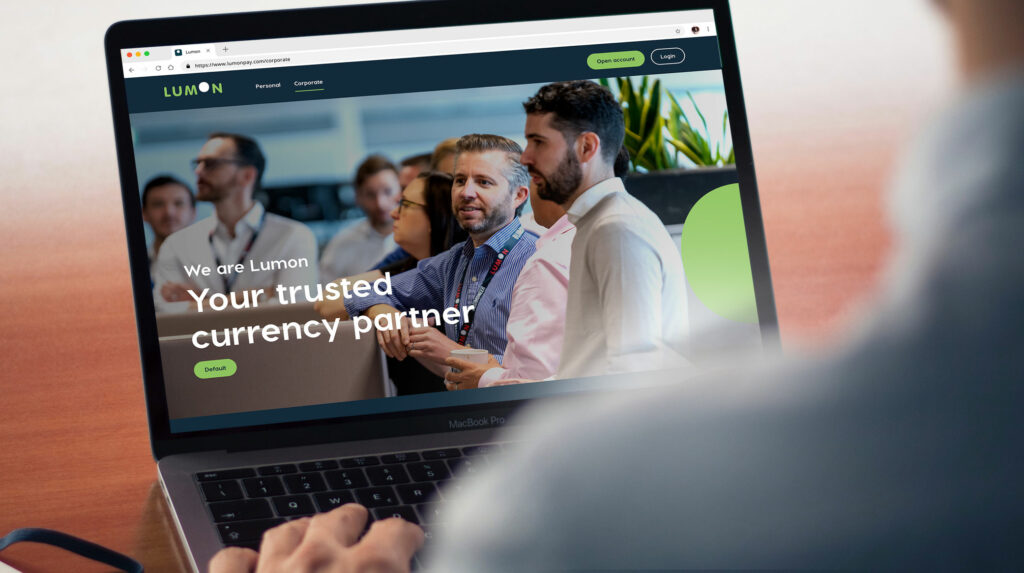 Over-the-shoulder view of a professional working on a MacBook Pro with a web page open to 'Lumon: Your trusted currency partner' featuring a team of engaged financial experts in the background.